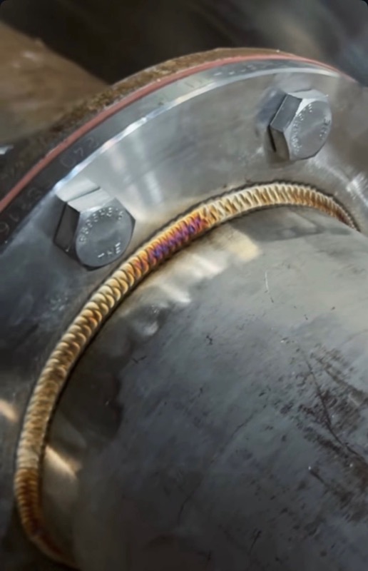 TIG bead curving around a stainless flange — gold heat tint, tight stack of dimes